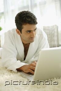 PictureIndia - Man in bathrobe, lying on floor at home, using laptop